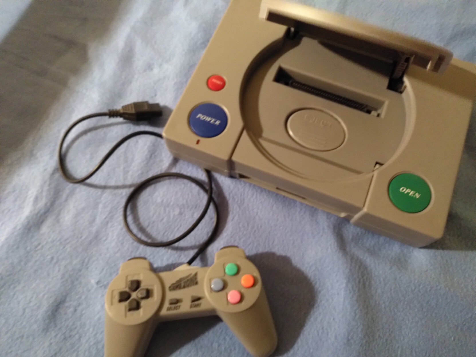 Photo d'une de mes famiclones, une console gris clair ressemblant fortement à une Playstation 1 mais avec des boutons colorés, l'emplacement pour placer le disque est remplacé par un emplacement cartouche, le pad ressemble aussi à un pad PS1 de l'époque.