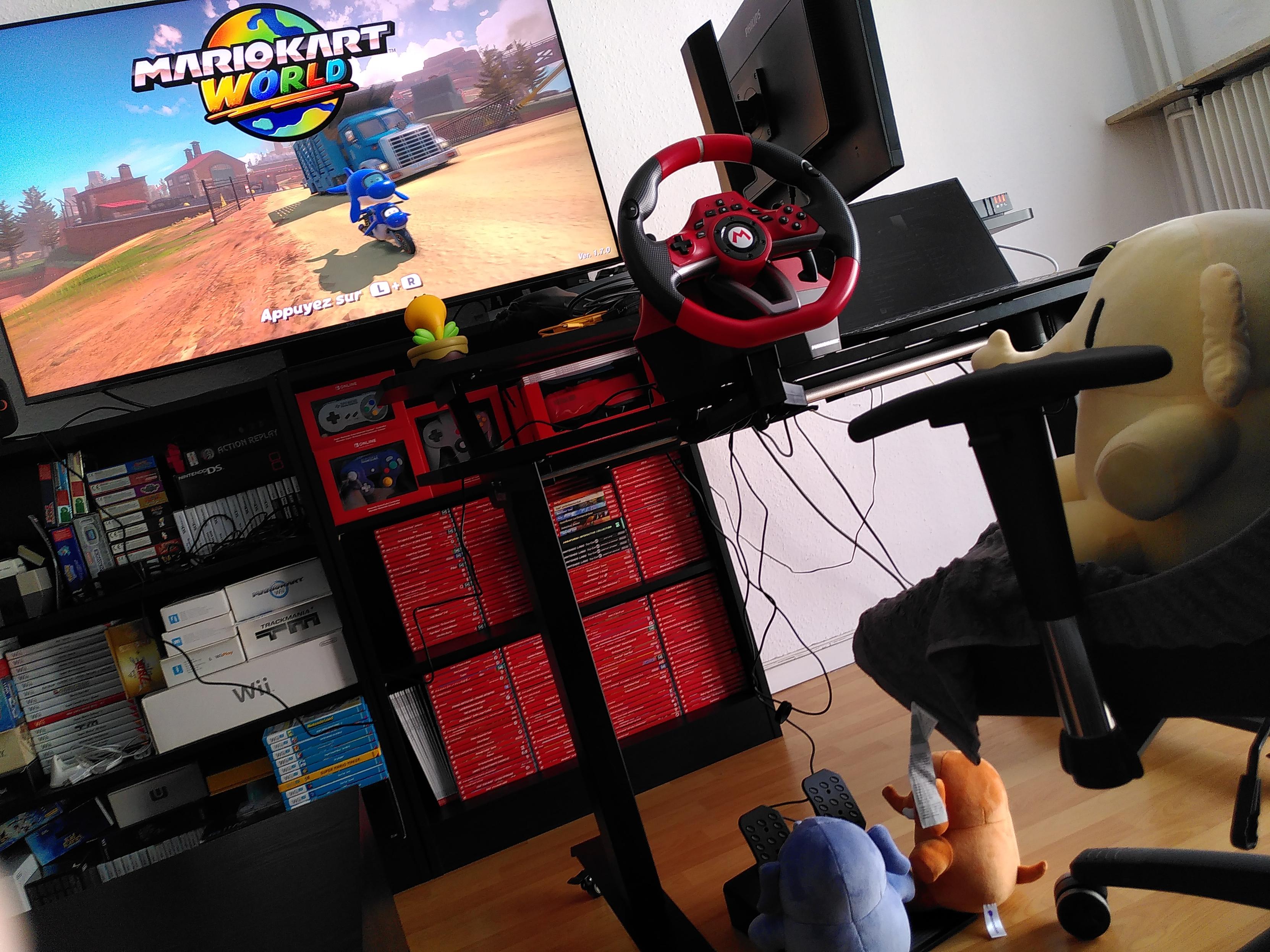 Photo de mon setup pour jouer à Mario Kart World : ma télé de salon affiche le jeu, le bureau mobile a été placé devant la télé pour que je puisse y visser un volant USB (et mon écran de PC a été pivoté pour laisser le champ de vision libre), des pédales sont reliées au volant et posées au sol, ma chaise de bureau est devant mon bureau. Plushtodon beige est assis sur ma chaise de bureau face au volant, Plushtodon Bleu et Pêche sont chacun en train d'appuyer sur une pédale au sol. En détails rigolos, on peut aussi noter la figurine Fleur Cancan pivotée pour qu'elle regarde l'écran, et le pilote affiché à l'écran est Dauphin sur le scooter Dauphin. Cocasse.