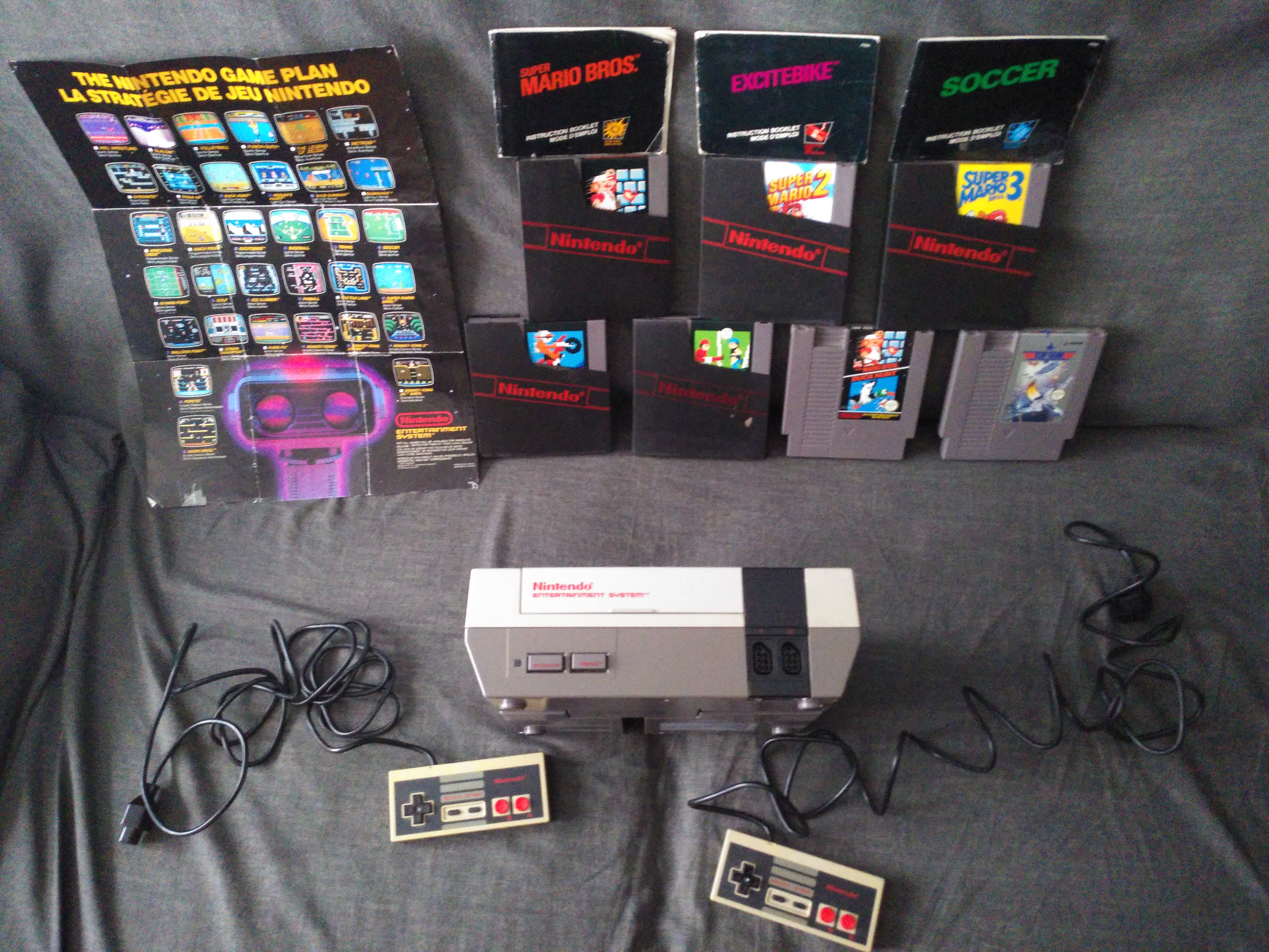 Photo de ma NES avec deux manettes jaunies, le poster vu plus au-dessus ; les cartouches des trois Super Mario Bros. occidentaux, d'Excitebike, de Soccer, du duo Super Mario Bros./Duck Hunt alors que j'avais pas de Zapper, et de Top Gun que j'ai jamais réussi à faire marcher ; j'ai encore cinq fourreaux mais 0 boite, et les modes d'emploi de Super Mario Bros., Excitebike et Soccer.